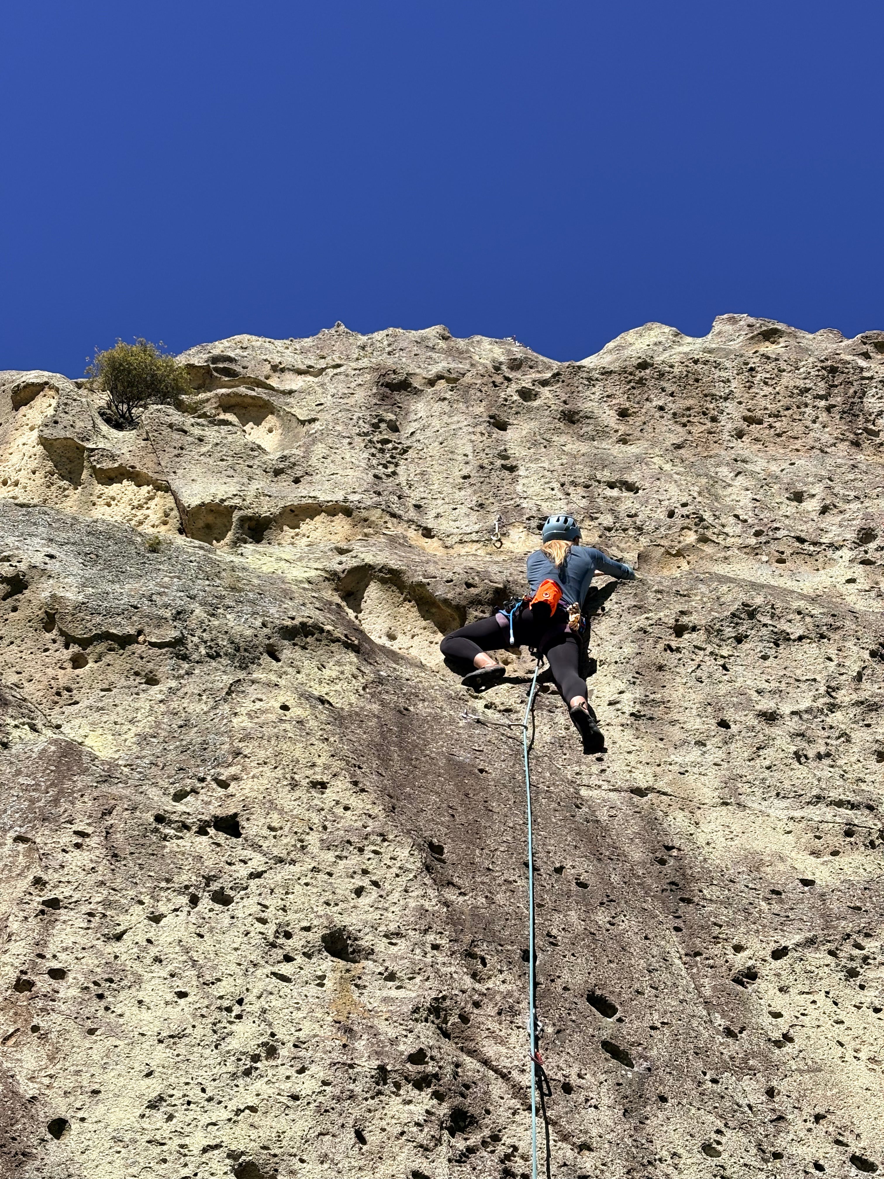 A person climbing a steep rocky cliff under a clear blue sky, wearing climbing gear and attached to a rope.