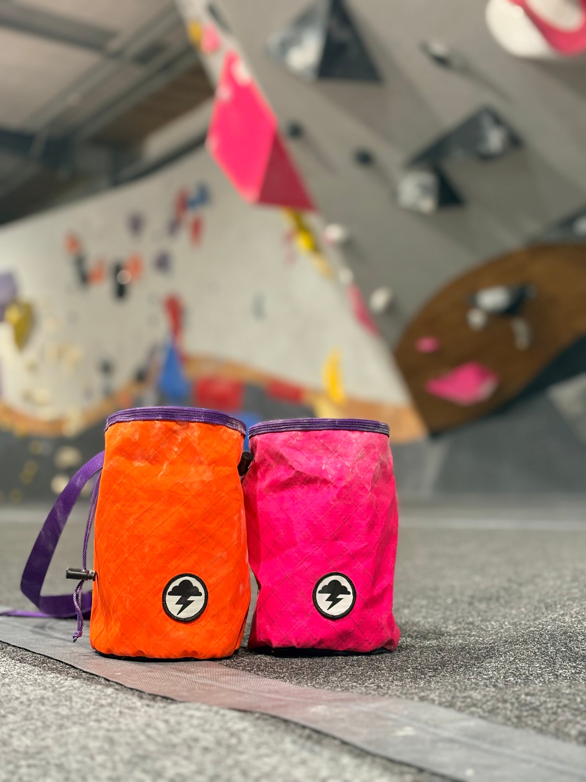 Two textured climbing chalk bags, one bright orange and one vivid pink, with purple straps and a circular logo patch, resting on a climbing gym floor with a blurred climbing wall in the background.